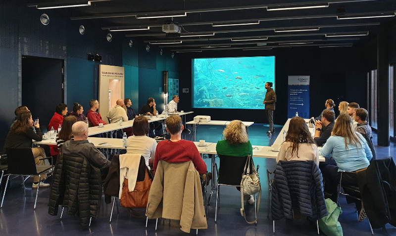 Gruppenaufnahme der Teilnehmer des 7. respACT Initiativkreises bei einem Vortrag im Konferenzraum des Alpenzoos Innsbruck vor einer Leinwand mit Unterwasser-Darstellung.