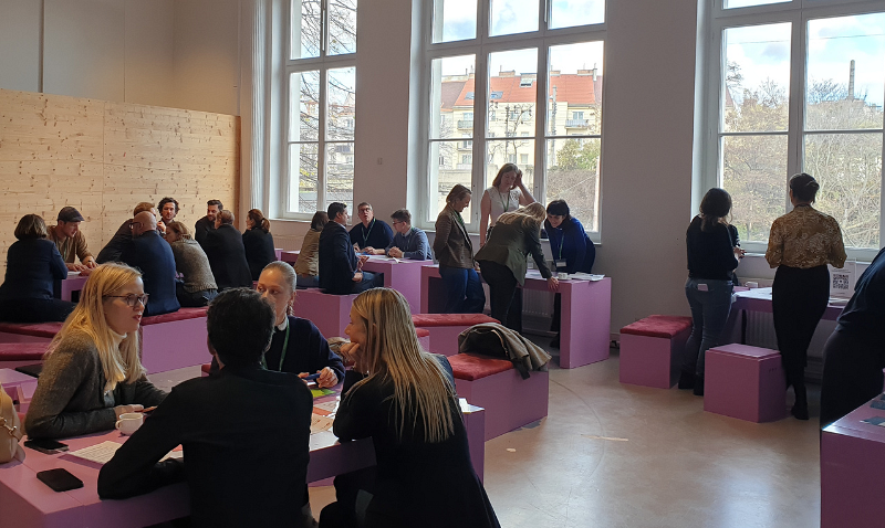 Menschen sitzen und stehen in einem hellen Raum mit großen Fenstern, versammelt um lila Tische und Hocker, während sie sich unterhalten und arbeiten.