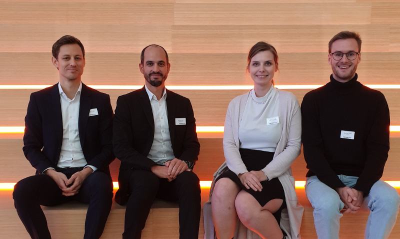 Vier Personen sitzen nebeneinander auf einer Treppe mit beleuchteten Stufen; zwei Männer tragen Anzüge, während die Frau und der vierte Mann in eleganter Kleidung gekleidet sind.